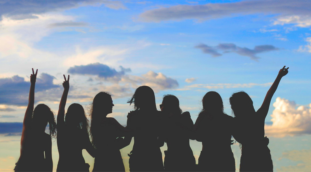 Silhouettes of seven women posing and throwing peace signs against a colorful sunset sky.