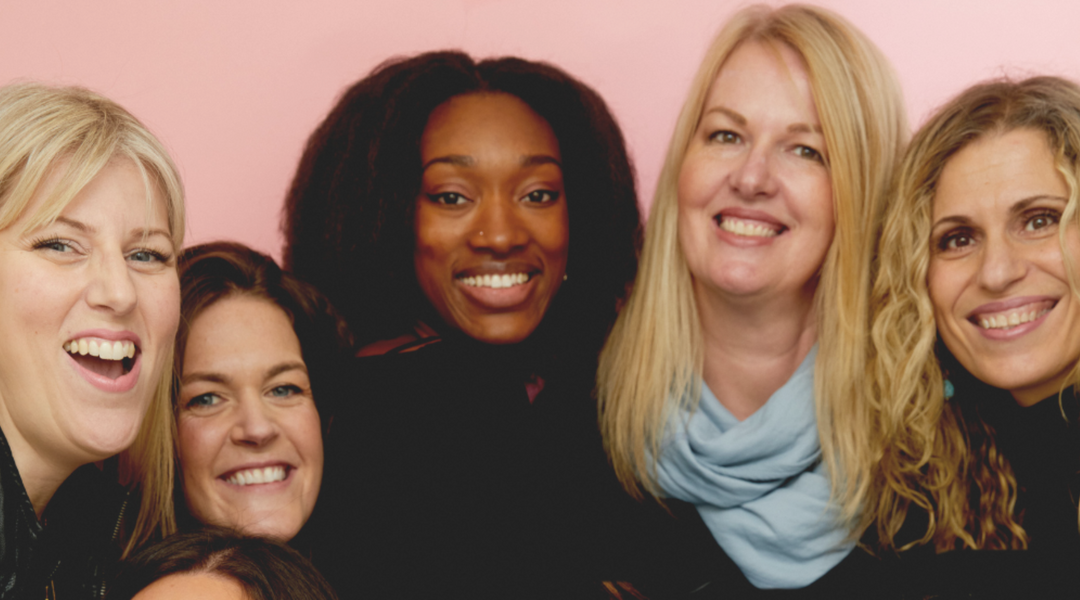 Tall women smiling and embracing in front of a pink backdrop.