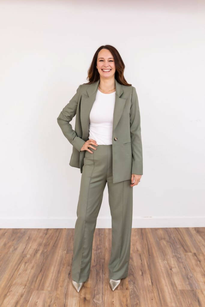 Tall woman wearing a sage green pantsuit with a white top and pointed silver heels, smiling confidently against a white backdrop.